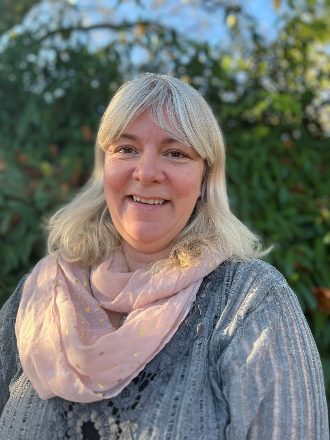 A woman with shoulder-length gray hair is smiling at the camera while standing outdoors. She is wearing a light pink scarf with small golden stars over a textured gray top. The background is filled with greenery, suggesting a natural, outdoor setting, and the lighting gives a warm, soft tone to the image.