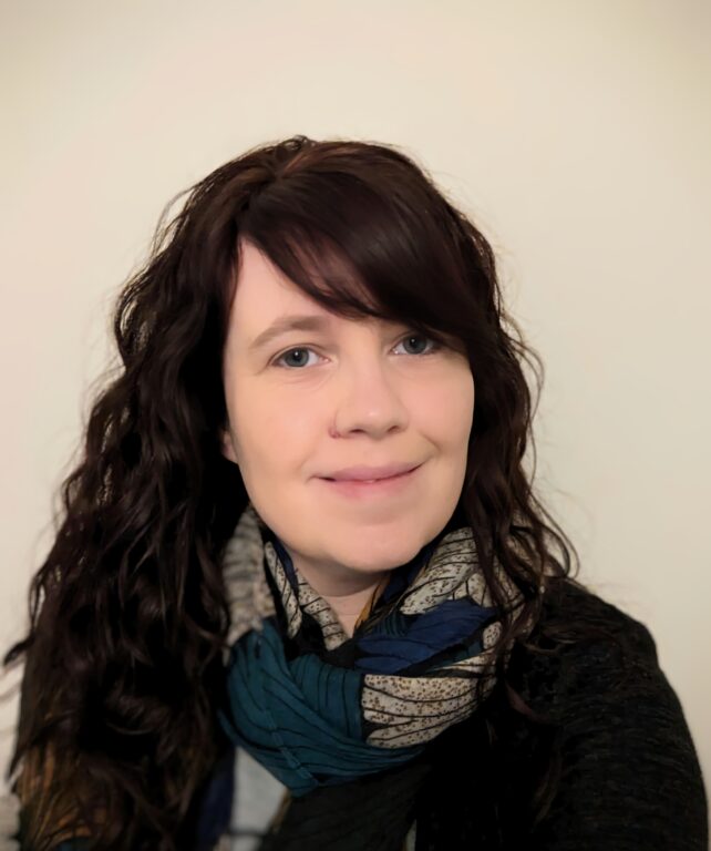 A woman with long, wavy dark hair is smiling gently at the camera. She is wearing a multicolored scarf with blue and beige tones over a dark outfit. The background is plain and light-colored, which brings focus to her face and expression.