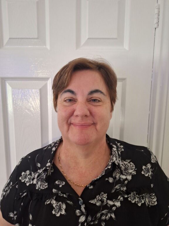 A woman with short brown hair is standing in front of a white paneled door, smiling at the camera. She is wearing a black blouse with a white floral pattern and a gold chain necklace. The background is simple, focusing attention on her warm and approachable expression.