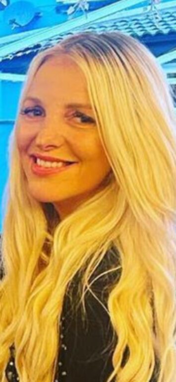 A close-up photo of a woman with long blonde hair, smiling warmly at the camera. She is wearing a black outfit, and her hair falls in loose waves over her shoulders. The background appears to be outdoors, with a blue-tinted structure or awning visible behind her, adding a soft, cool hue to the scene