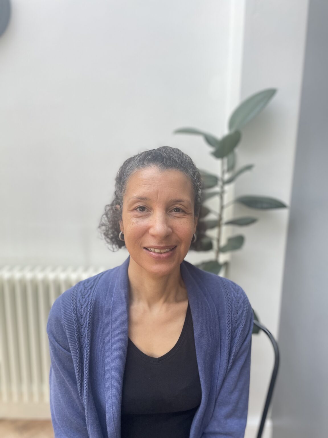 A woman with short, curly hair is sitting and smiling at the camera. She is wearing a dark top with a blue cardigan over it. The background is a softly lit indoor space with light-colored walls, a radiator, and a leafy green plant positioned behind her to the right. The overall atmosphere is calm and welcoming.