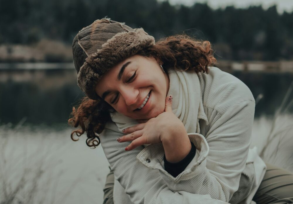 A young woman with curly hair is smiling warmly with her eyes closed, resting her chin on her hand in a relaxed and content pose. She is wearing a cozy, fur-lined hat and a thick scarf, along with a light-colored jacket, suggesting cold weather. The background features a serene outdoor setting with a calm body of water and blurred trees, creating a peaceful, nature-filled atmosphere.