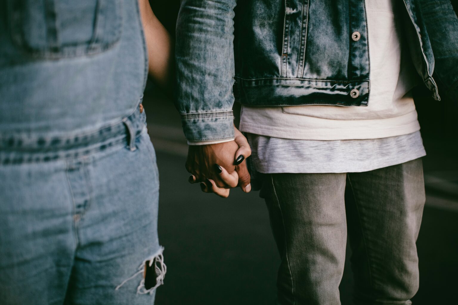 A close-up shot of two people holding hands while standing side by side. Both are wearing denim outfits: the person on the left is dressed in ripped jeans, while the person on the right is wearing a denim jacket over a layered shirt. The focus is on their clasped hands, emphasizing their connection and the casual, relaxed vibe of their attire.