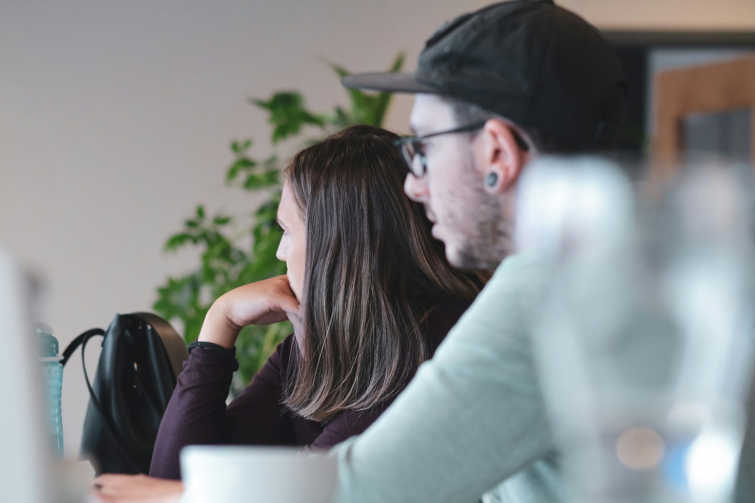 Two people are seated at a table, viewed from a side angle, focusing intently on something in front of them. The person on the left, with long brown hair, rests their chin on their hand, while the person on the right, wearing glasses and a cap, looks in the same direction. The background includes some out-of-focus green plants, adding a touch of nature to the indoor setting. The image has a soft, natural lighting, creating a relaxed and thoughtful atmosphere.