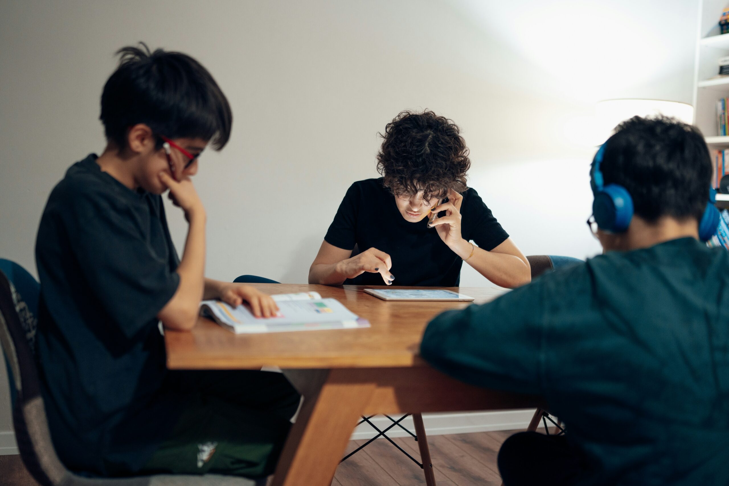 Three people are seated around a wooden table in a well-lit room, each engaged in different activities. The person on the left, wearing glasses, is focused on a book in front of them, resting their head on their hand in a thoughtful pose. The person in the center, with curly hair, is using a tablet while talking on a phone. The person on the right is wearing blue headphones and appears to be looking at something on the table, possibly a book or a tablet. The background features a white wall and a lit floor lamp, creating a cozy, focused atmosphere.