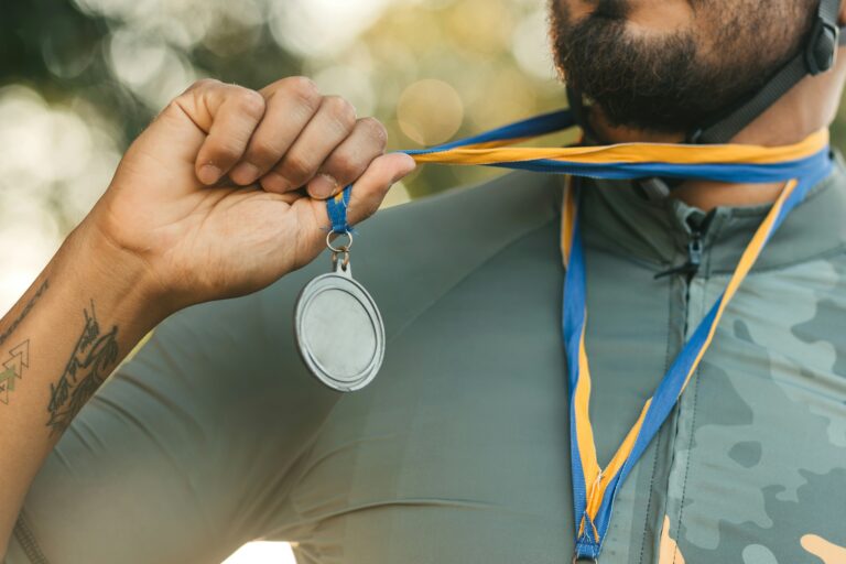 A close-up of a cyclist holding a silver medal by its blue and yellow ribbon, with a visible tattoo on their arm.