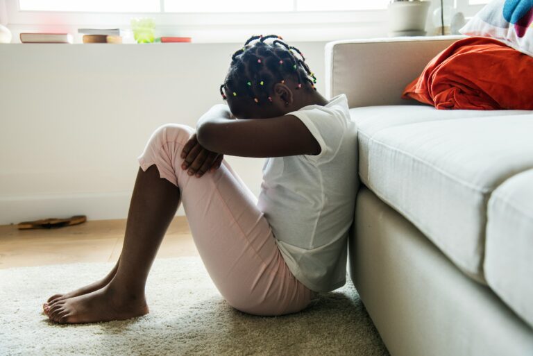 A young girl sits on the floor with her arms crossed over her knees and her head resting on them, appearing sad or reflective.