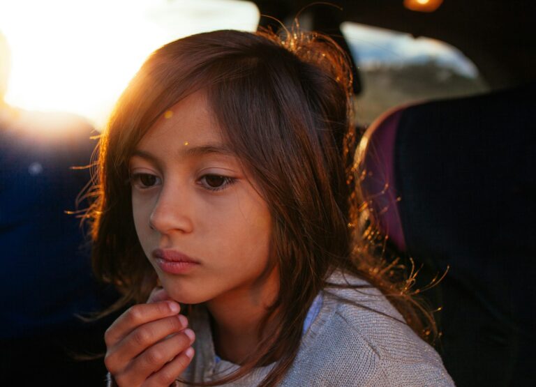 A young girl sitting in soft sunlight, looking thoughtful and introspective.