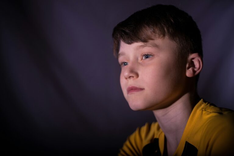 A young boy in a yellow shirt gazes thoughtfully into the distance, lit softly against a dark background.