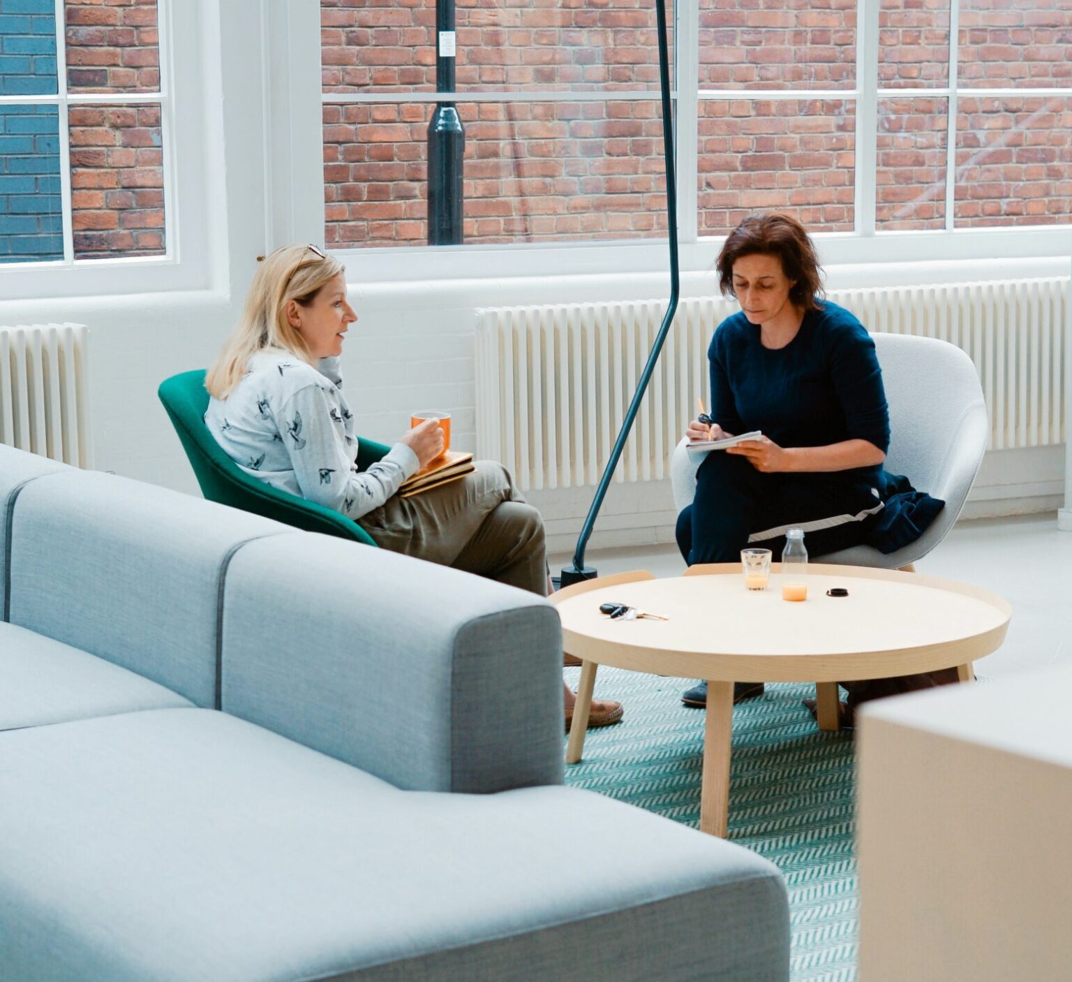 two women sat across from one another having a deep chat. One is note taking. They are in a co-working open space.