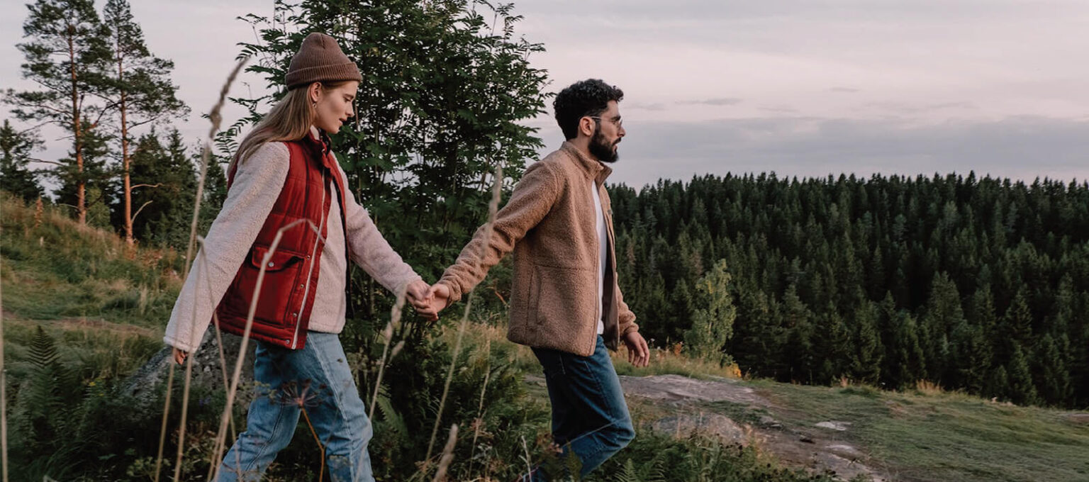 A couple holding hands, walking outdoors on a grassy hill with trees in the background. Both are dressed warmly in casual autumn attire