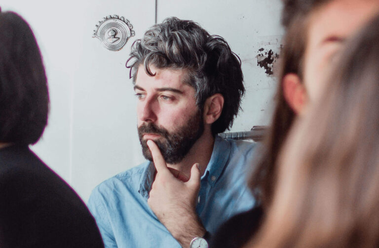 A man with a beard and curly hair, wearing a light blue shirt, is thoughtfully resting his hand on his chin. He appears to be deep in thought, with a focused expression on his face. The background shows blurred figures of other people, creating a sense of being in a group setting or discussion.