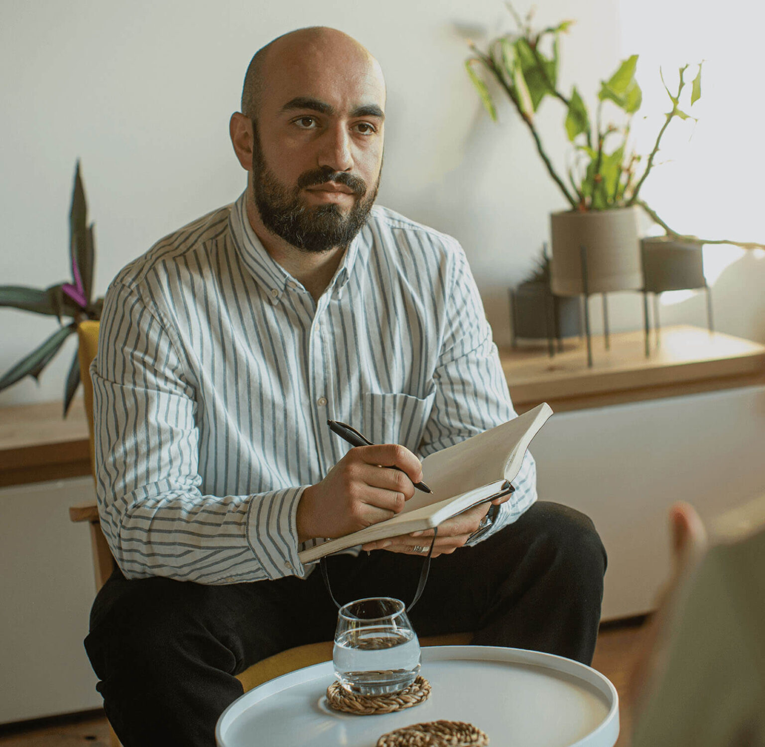 A bearded, bald man in a striped shirt writes in a notebook with a glass of water and plants in the background.