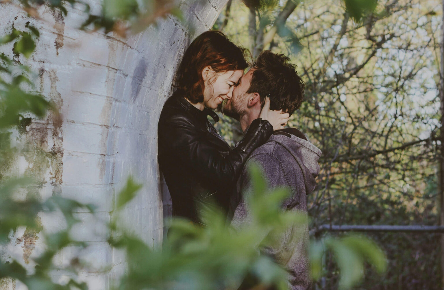 A couple sharing an intimate moment outdoors, with the woman leaning against a wall and the man close to her, both embracing affectionately amidst a backdrop of greenery.