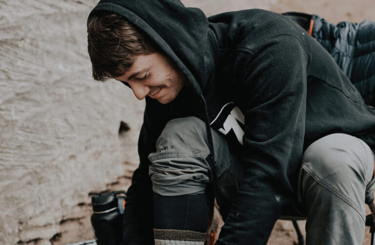 A young man with brown hair, wearing a black hoodie and grey pants, is kneeling down to tie his shoe.