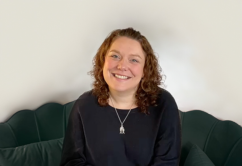 A woman with curly hair sits on a dark green sofa, smiling warmly at the camera. She wears a black top and a pendant necklace. The plain, light background keeps the focus on her friendly expression.