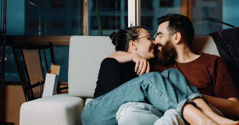 A couple sits closely on a couch, smiling with their noses touching in a warm, intimate moment. The cosy room has a large window with a night-time city view