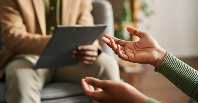 Person of colour with arms outstretched, as if in deep conversation. The hands are female and she wears a light green fine knit jumper with the sleeves in shot. Across from here is a male figure on a grey armchair, he is consulting a clipboard and is dressed very professionally in neutral toned smart blazer, shirt and trousers.