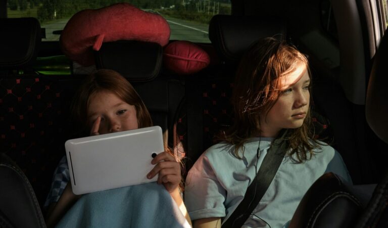 Two children are sitting in the back seat of a car; one is using a tablet, and the other is gazing out of the window.