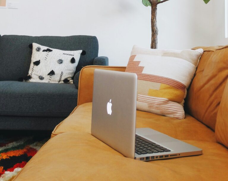 An open MacBook sits on a tan leather couch with a patterned pillow, while a dark blue armchair with a textured cushion is in the background