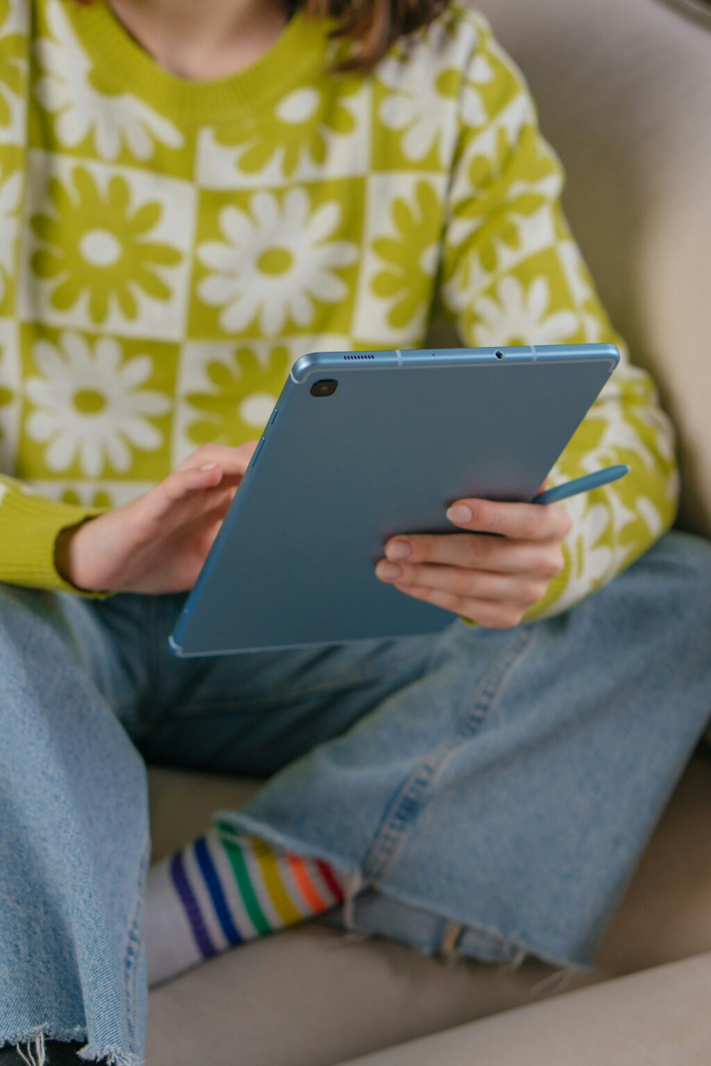 A person wearing a green daisy-patterned sweater and blue jeans sits on a couch, holding a blue tablet with a stylus