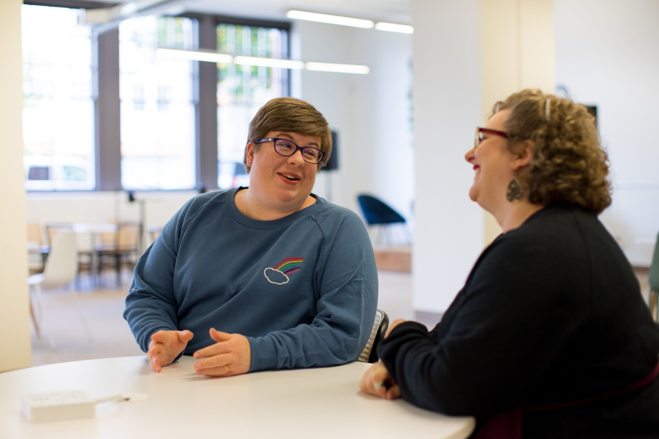 Two people sitting at a table in a bright indoor space, engaged in a friendly conversation and smiling.