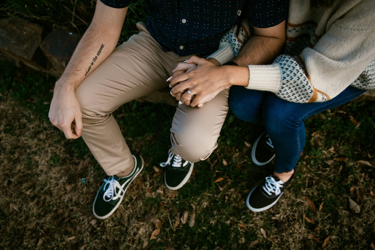 Two people hold hands. Their faces aren't visible. The person on the left wears light brown trousers and has a tattoo that says music is the answer. The person on the right wears jeans and wears a diamond ring on a well manicured hand.
