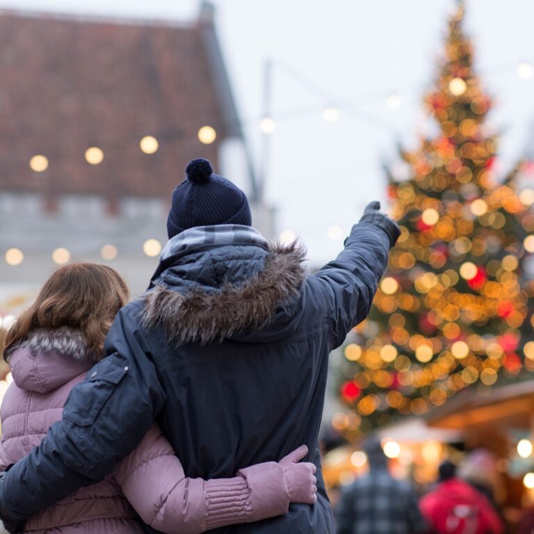 A couple of adults stand outside with their arms around each other's waist, looking in the same direction towards a Christmas tree. The person on the left wears a purple coat and the person on the right wears a navy coat and bobble hat, and is pointing towards the Christmas tree.