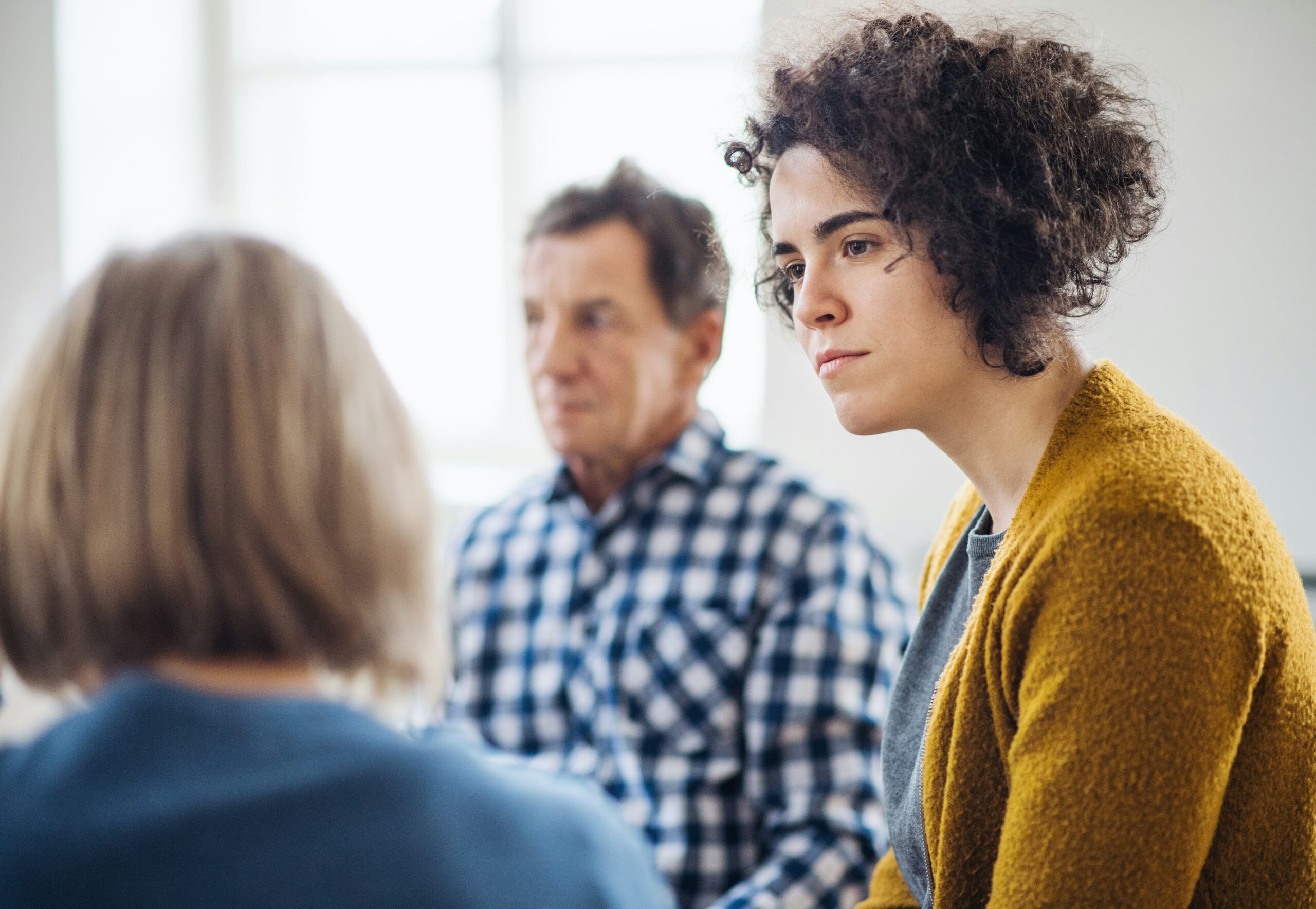 2 adults sit for therapy. In the foreground a therapist and 20's something woman. In the background a figure which appears to be her dad in age