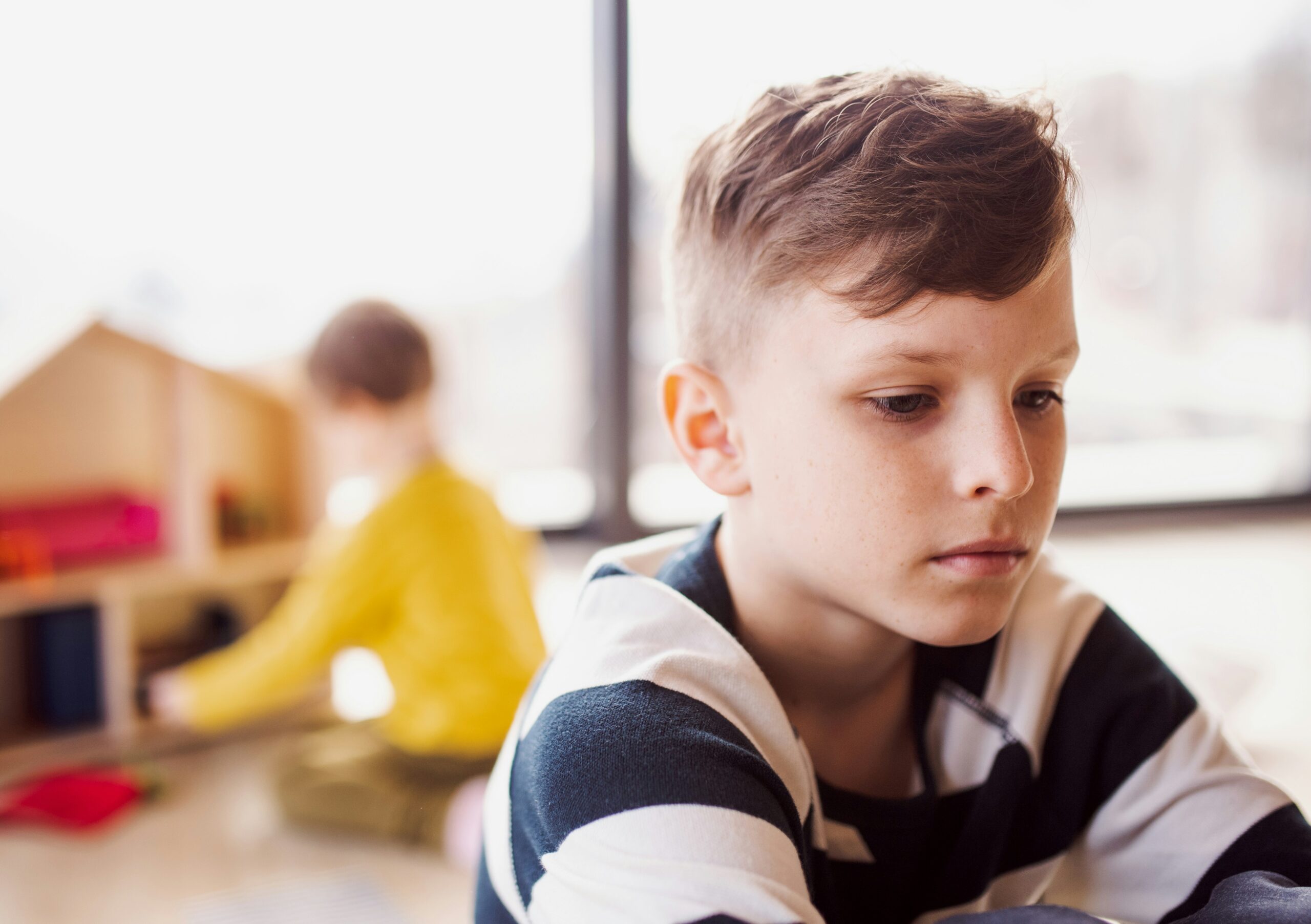 A child looks serious and has his back turned to another child who is playing in the background.