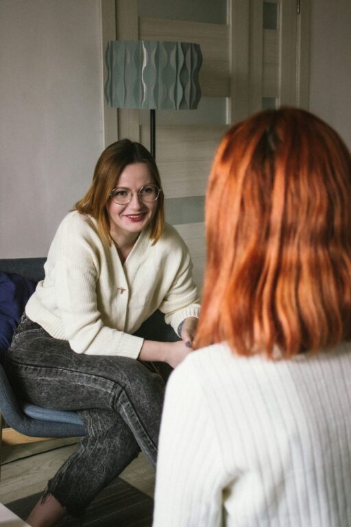 Two people sit facing each other in conversation. The one facing the camera wears glasses and is smiling. The one with their back to the camera has red shoulder-length hair. 