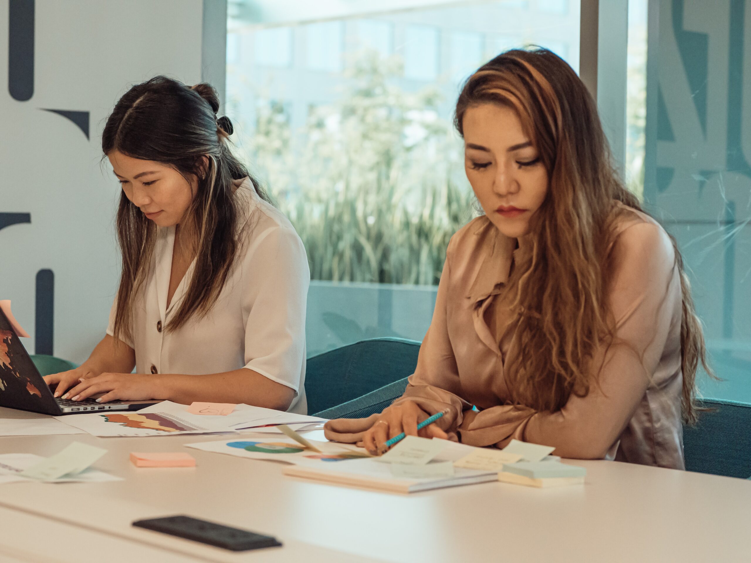 Two women in a modern office space work at a table, one using a laptop and the other writing on sticky notes.