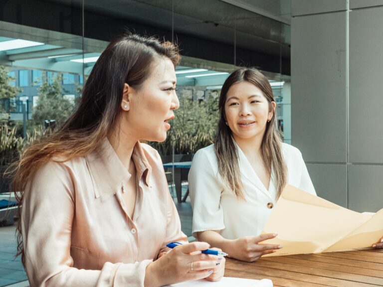 Two women sitting outside at a table, engaged in a conversation, with one holding a coffee cup and the other holding a folder.