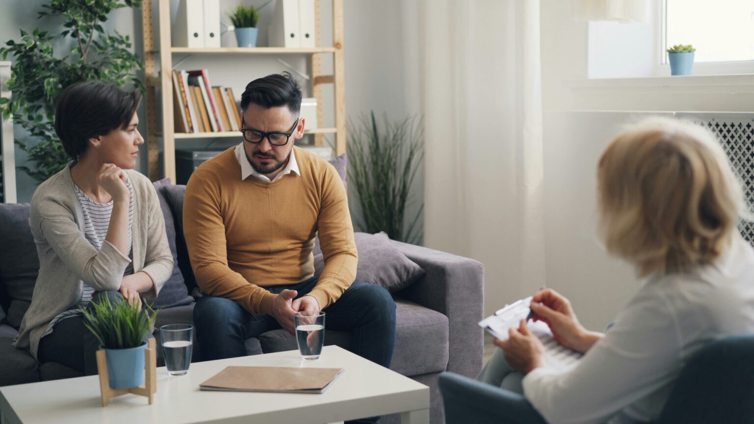 Two people share a sofa and talks with a third person who faces them, holding a clipboard.