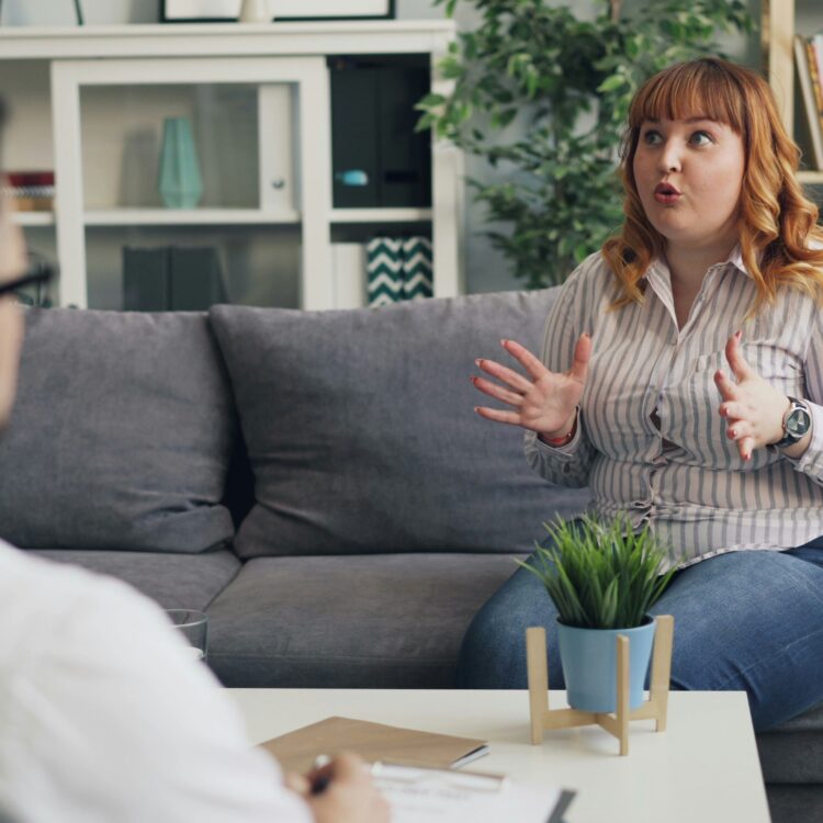 Two people face one another while sitting on couches. The one facing the camera appears to be speaking animatedly while the other listens attentively.
